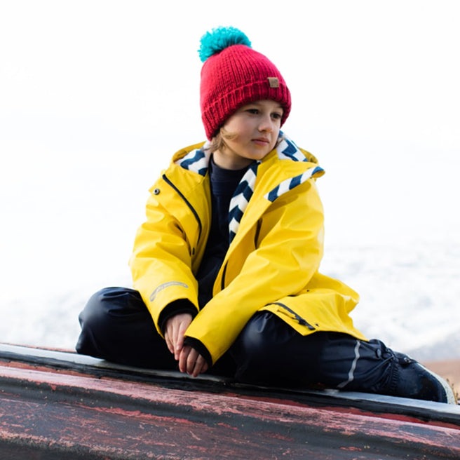 A boy in a Puddleflex Waterproof Fleece Lined Jacket in yellow, sitting on a boat wearing a red hat with a blue pom pom.